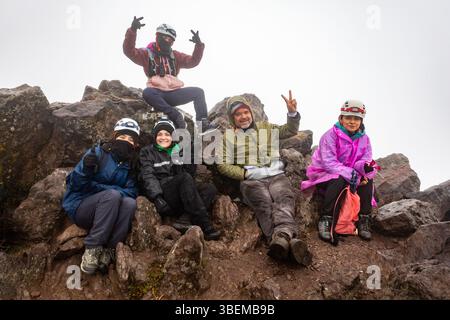 Groupe de randonneurs en vêtements de pluie colorés et casques prennent une pause sur un terrain rocheux pendant une journée de pluie. Ils s'assoient et posent avec joie et enthousiasme. Banque D'Images