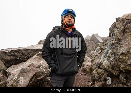 Un randonneur déterminé portant un équipement de pluie et un casque de sécurité se tient en toute confiance au milieu d'un sentier de montagne rocheux. L'atmosphère brumeuse renforce l'aventuro Banque D'Images