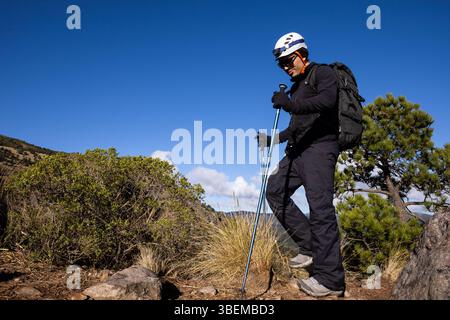 Un randonneur portant un équipement de plein air et un casque marche avec des bâtons de trekking sur un sentier de montagne rocheux, entouré de verdure et d'un ciel bleu clair. Aventure a Banque D'Images