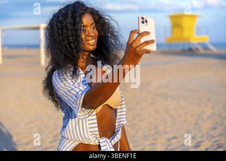 Jeune femme noire prenant selfie sur la plage à l'aide d'un smartphone, portant une chemise rayée et un haut de bikini, avec tour de sauveteur en arrière-plan Banque D'Images