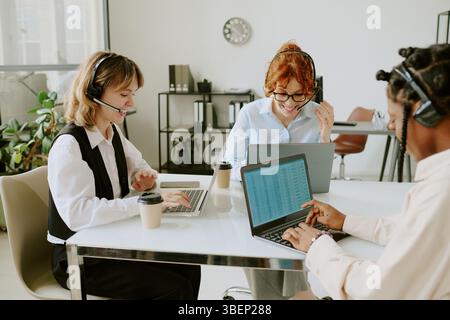 Équipe diversifiée travaillant sur des ordinateurs portables et s'engageant dans des discussions animées dans un environnement de bureau moderne. Micro-casques et tasses à café contribuant à une atmosphère de travail intense Banque D'Images