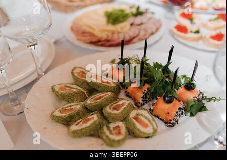 Cadre de table élégant avec une variété de hors-d'œuvre, y compris des rouleaux de sushi et des assiettes de viande, disposés sur une nappe blanche, avec des verres à vin et Banque D'Images