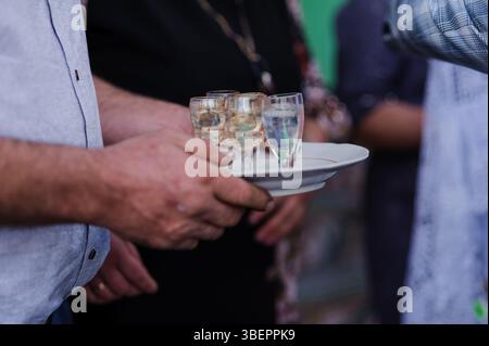 Gros plan capturant les mains d'un serveur tenant un plateau de trois petits verres remplis de liquide clair, probablement des boissons alcoolisées, pendant un événement Banque D'Images