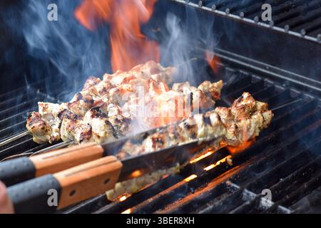 Gros plan pinces rotatives brochettes de poulet grillant sur un barbecue avec du feu et de la fumée Banque D'Images