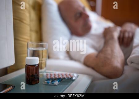 Homme senior dormant paisiblement dans le lit avec des pilules et un verre d'eau à côté de lui sur la table de nuit dans sa chambre. Focus sélectif sur bouteille de pilules. Banque D'Images