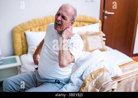 Homme âgé ayant mal au cou, il crie alors qu'il est assis sur le lit après s'être réveillé tôt le matin. Banque D'Images