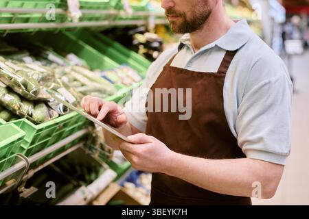 Homme parcourant la tablette tout en se tenant devant la section des produits frais à l'épicerie portant un tablier, se concentrant sur la gestion des stocks Banque D'Images