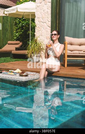 Une femme brune des années 20 en maillot de bain se détend au bord de sa piscine privée tout en profitant d’un petit déjeuner flottant dans l’eau. Banque D'Images