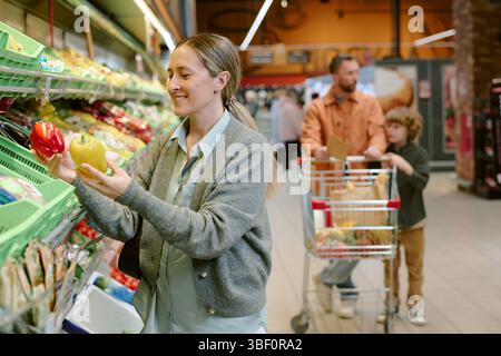 Femme examinant soigneusement les poivrons dans la section des produits de l'épicerie tandis que l'homme et l'enfant avec le chariot d'achat en arrière-plan Banque D'Images