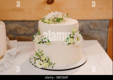 Un gâteau de mariage blanc à deux niveaux élégamment décoré avec un glaçage texturé et accentué de petites grappes de fleurs blanches et de fol vert frais Banque D'Images