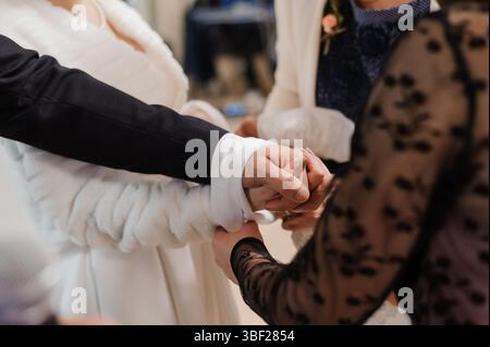 Cette photographie intime se concentre sur les mains serrées d'une mariée et d'un marié lors de leur cérémonie de mariage, avec des vêtements élégants et une grotte arrière floue Banque D'Images
