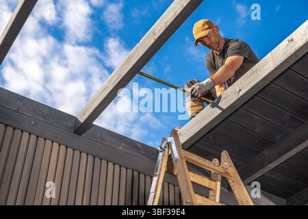 Un ouvrier de la construction se concentre sur la mesure des poutres en bois tandis qu'il est perché sur une échelle sous un ciel bleu vif. Il travaille sur un projet de rénovation. Banque D'Images