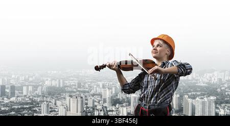 Belle jeune femme dans un casque de sécurité jouant du violon. Constructeur féminin professionnel souriant en chemise bleue à carreaux sur fond de paysage urbain. Industriel Banque D'Images