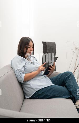 Une jeune étudiante en tenue semi-formelle est assise sur un fauteuil moderne, examinant ses notes sur une tablette avant d'entrer en classe. Le calme, le mépris Banque D'Images