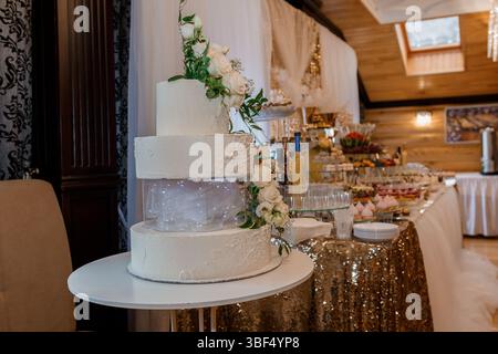 Un gâteau de mariage blanc magnifiquement décoré à trois niveaux avec de délicates arrangements floraux se trouve sur une table ronde blanche drapée d'un se doré scintillant Banque D'Images