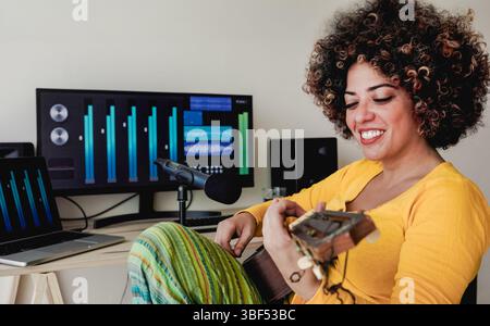 Jeune femme latina avec des cheveux afro, jouant ukulélé et souriant dans un studio d'enregistrement à domicile confortable - concept de technologie musicale Banque D'Images
