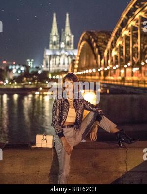 Jeune femme assise avec une jambe en haut devant la vue nocturne de Cologne en Allemagne. Pont éclairé appelé ''Die Hohenzollernbrücke'' Banque D'Images
