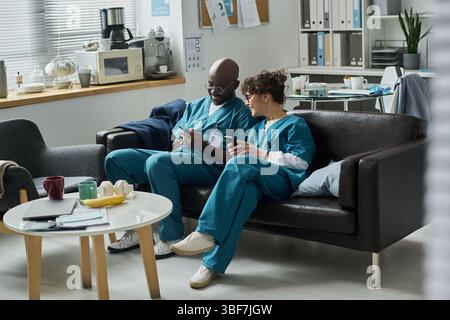 Professionnels de la santé assis sur le canapé et discutant des informations patient sur tablette. Étagères et équipement médical en arrière-plan avec des articles sur la table Banque D'Images