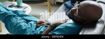 Professionnel de la santé reposant sur une chaise en cuir avec les mains pliées, portant des gommages bleus et des lunettes, paraissant calme et détendu en pause, mettant en valeur un moment décontracté dans un environnement occupé Banque D'Images