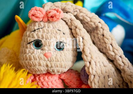 Un joli jouet de lapin en peluche avec texture de tissu présente de grands yeux bleus et un noeud rose. Il est placé parmi d'autres animaux en peluche colorés dans un environnement confortable Banque D'Images