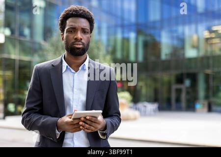 Un bel homme d'affaires noir se tient dehors, tenant une tablette devant un bâtiment moderne. L'accent est mis sur son expression sérieuse. Banque D'Images