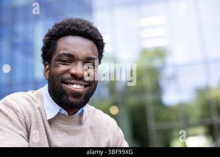 Un homme noir joyeux avec un sourire chaleureux est au centre de cette image, prise en extérieur avec un fond moderne. Il a l'air heureux. Banque D'Images