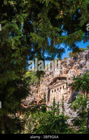 Anciennes tombes rupestres lyciennes sculptées dans une falaise à Kaunos, Dalyan, Turquie, partiellement encadrées par des arbres verts denses sous un ciel bleu. Banque D'Images