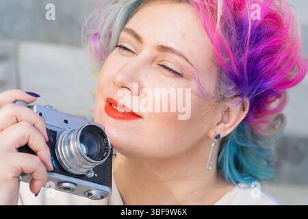 La femme caucasienne avec des cheveux de couleur curly prend des photos sur un ancien appareil photo rétro extérieur Banque D'Images