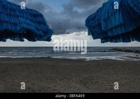 Deux parasols bleus se dressent sur une plage vide à la fin de l'été, avec un ciel orageux et une mer sombre en arrière-plan, créant une atmosphère mélancolique Banque D'Images