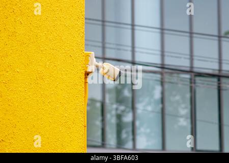 Une vue rapprochée d'une caméra de surveillance montée sur un mur jaune vif, juxtaposée contre un bâtiment en verre flou, mettant en valeur la technologie de sécurité moderne tec Banque D'Images