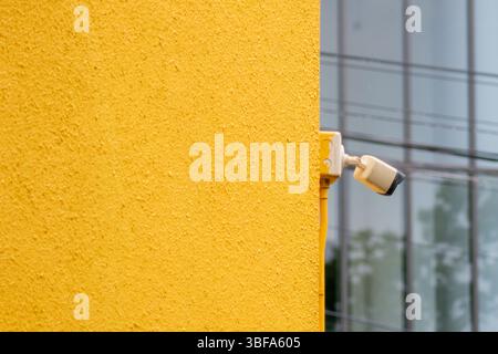 Gros plan d'une caméra de sécurité montée sur un mur jaune vif, contrastant avec un bâtiment moderne en verre en arrière-plan, symbolisant les s urbains Banque D'Images