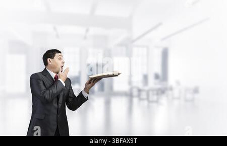 Peur d'affaires couvrant la bouche avec la main et tenant un livre ouvert. Surpris l'homme dans les affaires adultes costume et cravate sur fond de bureau permanent floue. Banque D'Images