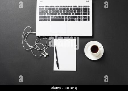 Nature morte du lieu de travail de bureau avec ordinateur portable et tasse de café. Surface noire plate avec écouteurs et bloc-notes vierge ouvert. Technologie numérique et m Banque D'Images