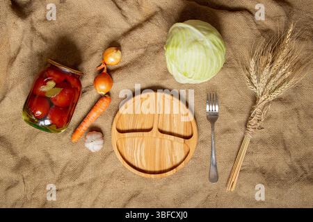 Légumes, chou, ail, carottes, oignons, blé et un pot de tomates en conserve sur toile de jute avec une assiette en bois et une fourchette, nourriture maison Banque D'Images