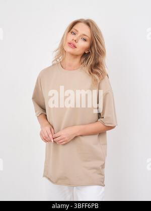 Femme en chemise beige oversize avec coupe décontractée, mains attachées devant, pantalon blanc. Cheveux blonds longs, expression neutre, maquillage naturel. Décontracté Banque D'Images