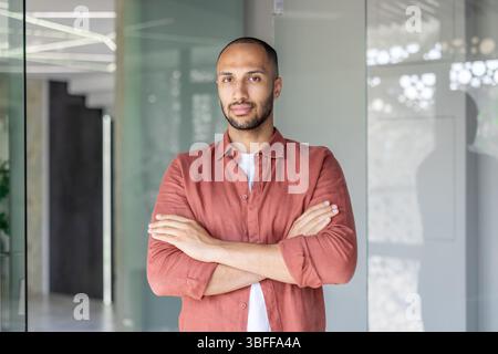 Un homme confiant aux bras croisés pose dans un cadre moderne, exsudant force et assurance. La lumière naturelle renforce l'atmosphère détendue. Banque D'Images