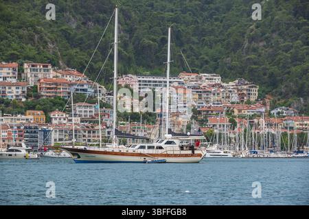 De nombreux bateaux, dont un yacht marron et blanc de premier plan, reposent dans les eaux portuaires de Fethiye, Türkiye, sur fond de bâtiments à flanc de colline. Banque D'Images