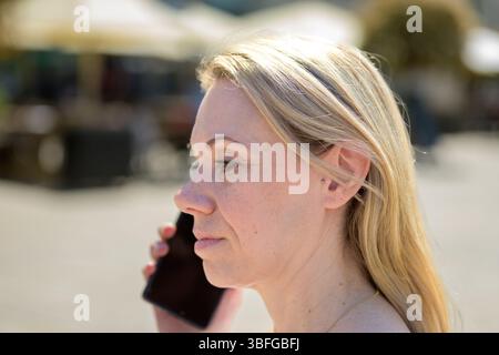Profil latéral d'une femme blonde parlant sur un smartphone en plein jour. Son expression semble concentrée et calme. Banque D'Images