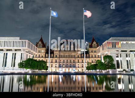 Vue nocturne du Capitole de l'État de New York à Albany, encadré par des bâtiments gouvernementaux modernes et des mâts de drapeau, avec des reflets spectaculaires sur l'eau calme de l'Empire State Plaza sous un ciel nuageux. Banque D'Images