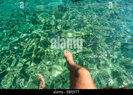Vue de dessus des jambes d’un homme jouant avec les eaux turquoises cristallines de la plage d’Agia Theodoti à iOS en Grèce Banque D'Images