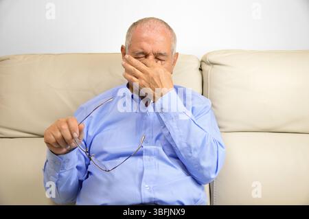 Homme âgé avec des lunettes souffrant de fatigue oculaire assis sur un canapé à la maison Banque D'Images