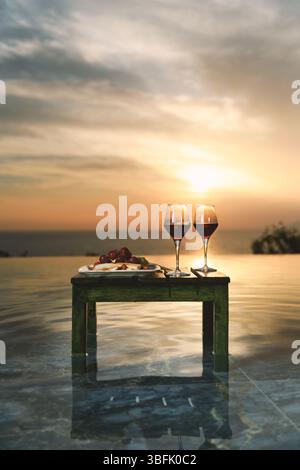 Deux verres de vin rouge et un élégant plateau de fruits présentés par une piscine à débordement au coucher du soleil. Banque D'Images