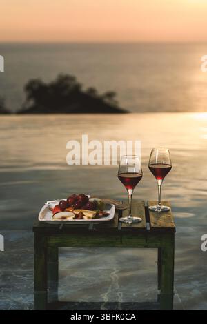 Deux verres de vin rouge et un élégant plateau de fruits présentés par une piscine à débordement au coucher du soleil. Banque D'Images
