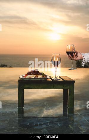 Deux verres de vin rouge et un élégant plateau de fruits présentés par une piscine à débordement au coucher du soleil. Banque D'Images