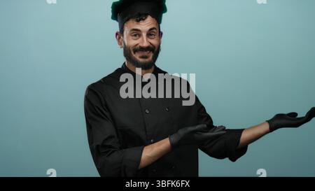 Chef hispanique en uniforme noir et gants présentant les mains ouvertes sur un fond bleu, souriant avec confiance dans un contexte culinaire. Banque D'Images