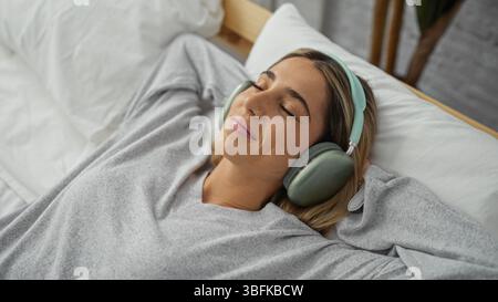 Femme se relaxant sur le lit portant des écouteurs dans un cadre de chambre confortable, exsudant le confort et la tranquillité avec les yeux fermés, profitant d'un moment paisible à ho Banque D'Images