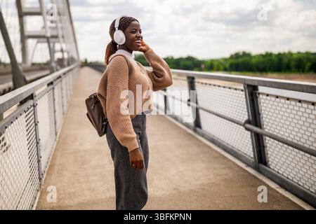 Heureuse jeune femme noire marche sur le chemin de pied de pont, écoute de la musique et sourit. Elle profite d'une journée ensoleillée en plein air. Banque D'Images
