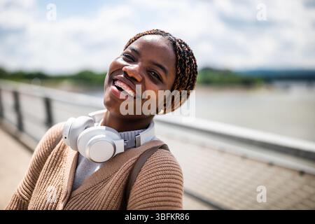 Heureuse jeune femme noire regarde la caméra et sourit. Elle profite d'une journée ensoleillée en plein air. Banque D'Images
