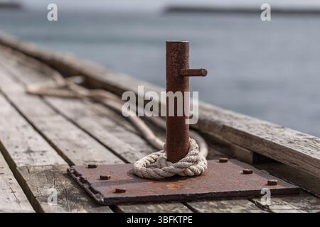 Borne d'amarrage en métal rouillé avec une vieille corde sur la jetée en bois altéré sur la côte des Lofoten, dans le nord de la Norvège. La mer et la jetée s'estompent dans un flou d'arrière-plan doux. Banque D'Images