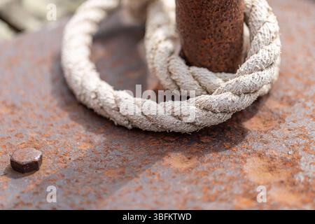 Gros plan d'une corde blanchie au soleil enroulée autour d'une borne en métal rouillé. La texture de la corde et les détails de corrosion sont clairement visibles. Banque D'Images